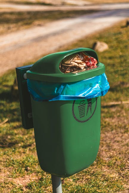 A green outdoor waste bin with a rounded lid, partially covered with a blue plastic bag, is situated on a grassy area during daylight. The bin has a black and white recycling symbol indicating waste s